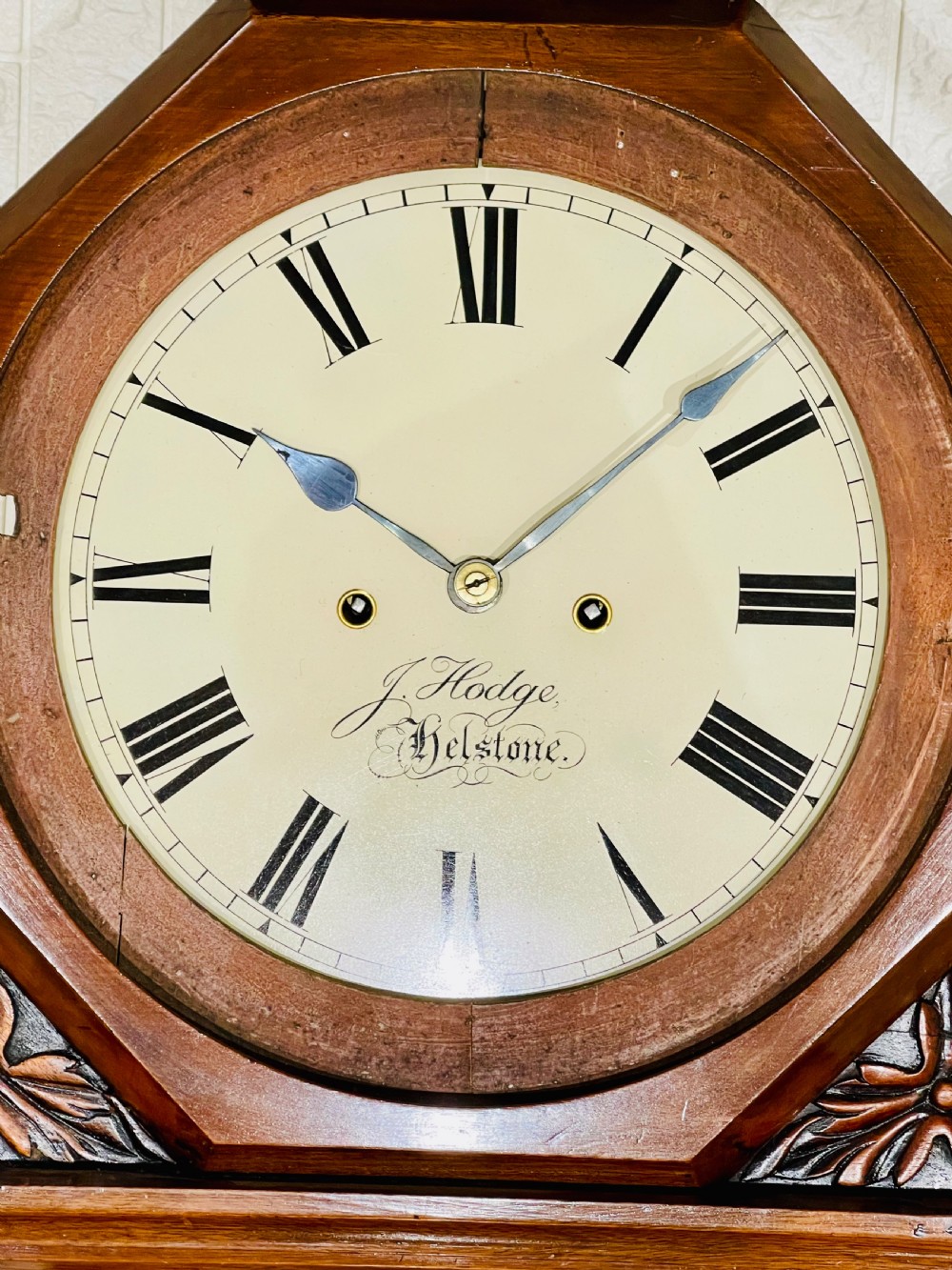 An antique Regency mahogany wall clock featuring an octagonal dial frame, carved floral shoulders, and a long trunk door with a key tassel. View VI