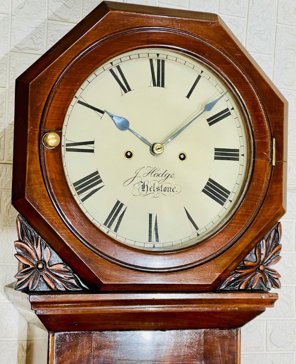 An antique Regency mahogany wall clock featuring an octagonal dial frame, carved floral shoulders, and a long trunk door with a key tassel. View VII