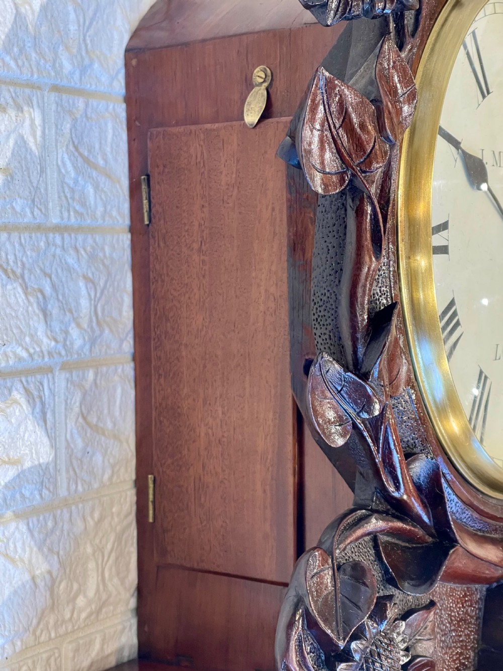 A Victorian carved mahogany fusee wall clock with Roman numerals on a white dial framed by carved floral details, resting on a matching wooden wall bracket. View VII