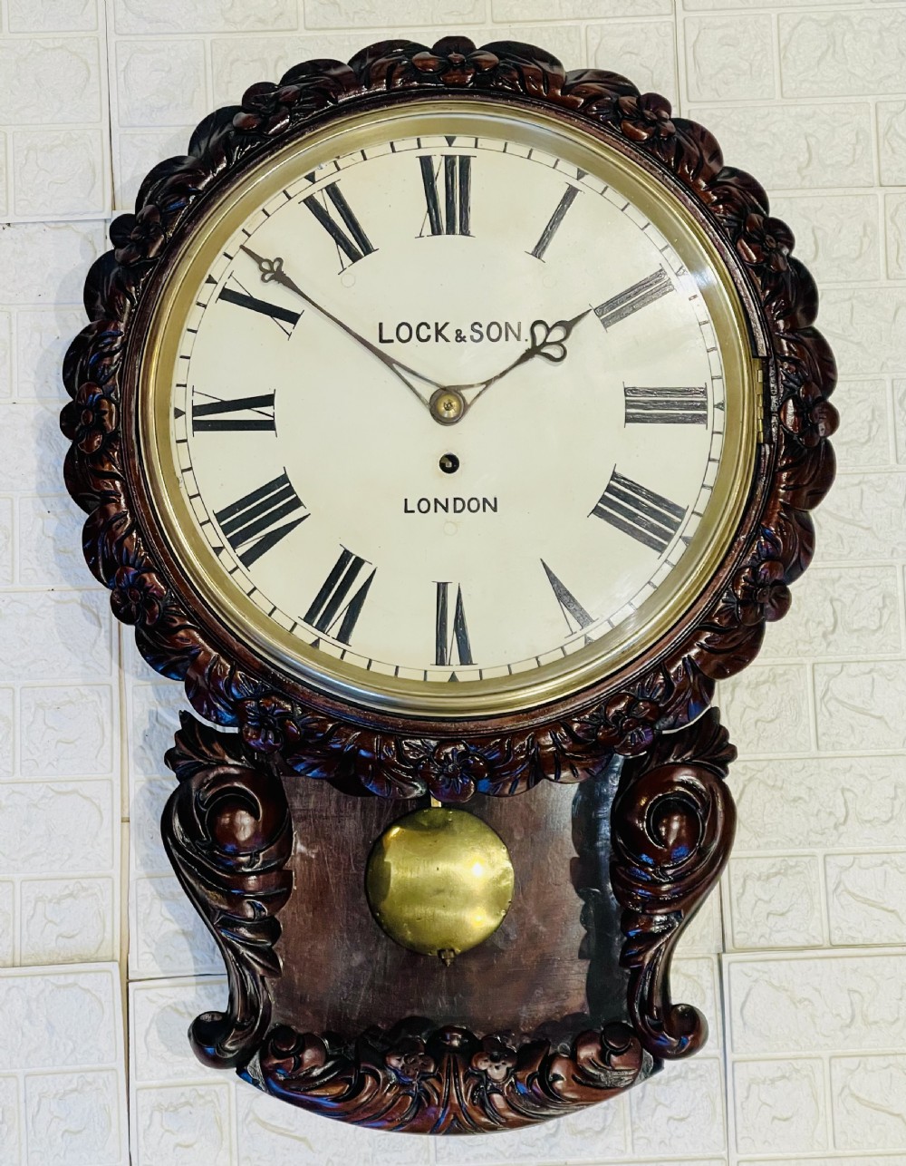 An antique dark mahogany drop dial wall clock featuring a white dial face with Roman numerals and a visible brass pendulum.