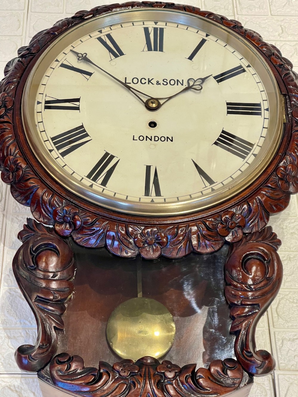 An antique dark mahogany drop dial wall clock featuring a white dial face with Roman numerals and a visible brass pendulum. View V