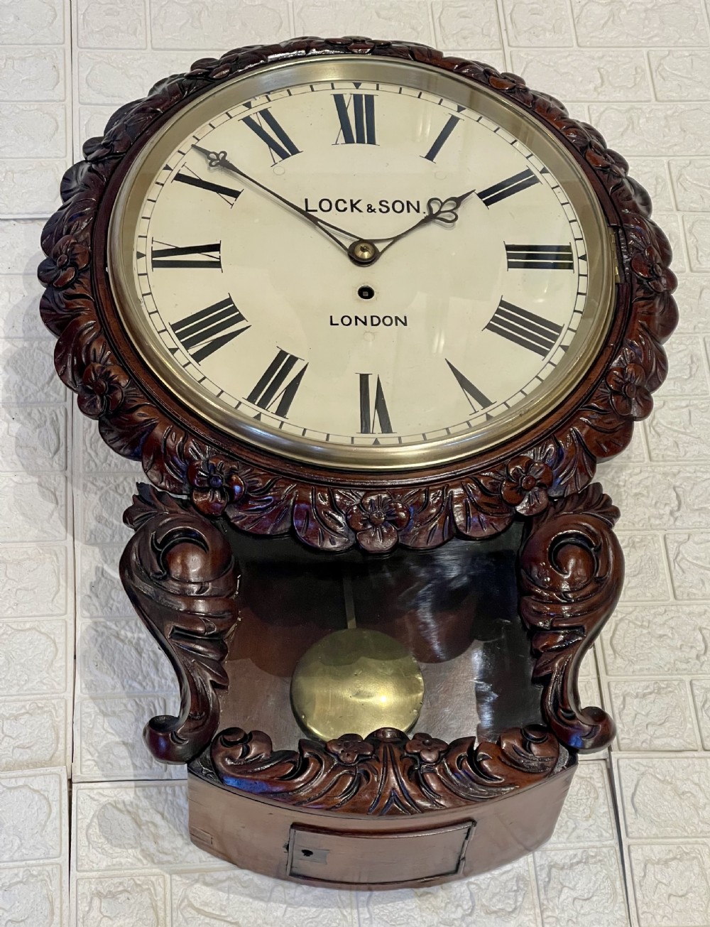 An antique dark mahogany drop dial wall clock featuring a white dial face with Roman numerals and a visible brass pendulum. View VI