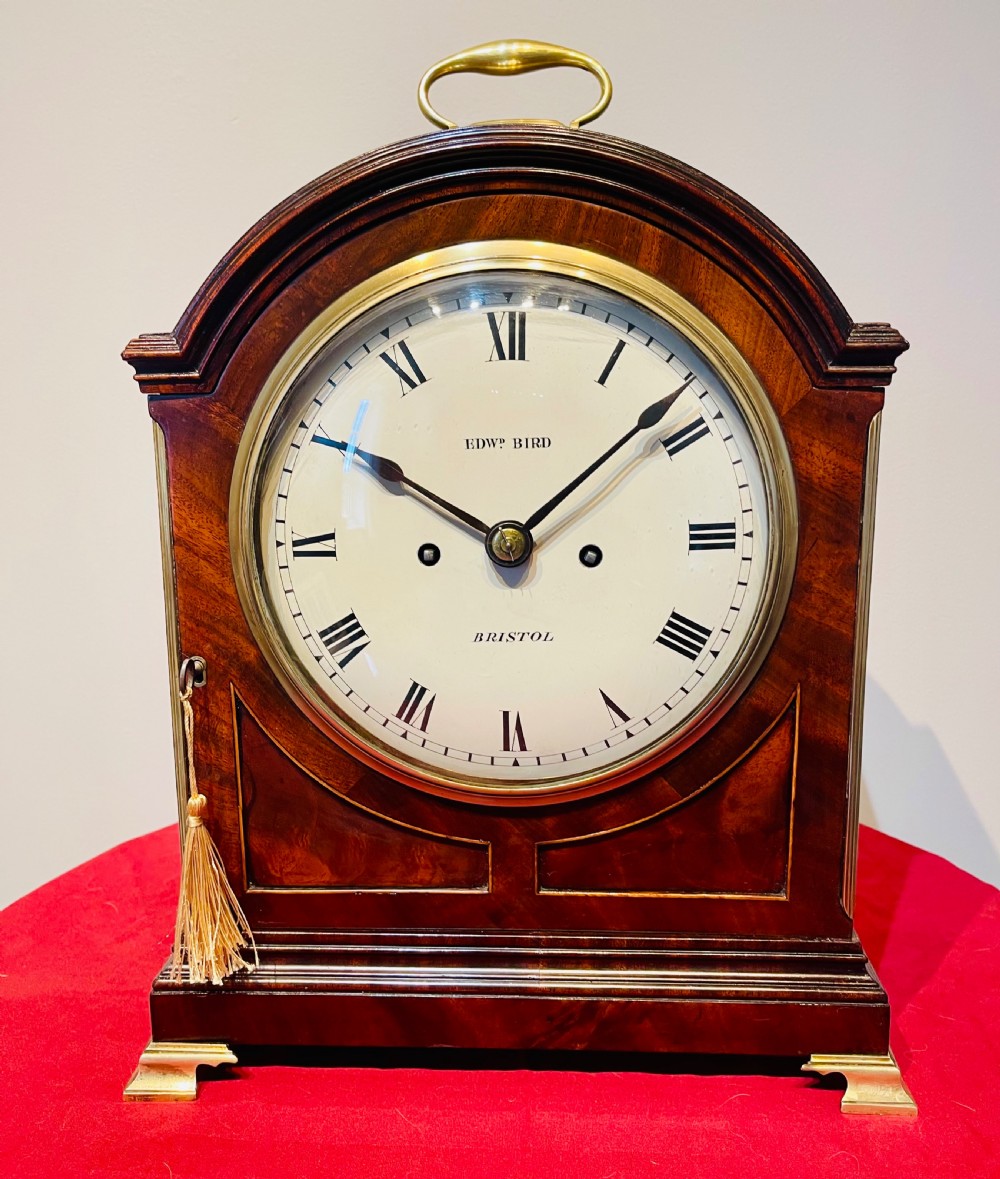 A George III mahogany bracket clock featuring an arched top with a brass handle, a circular white dial with Roman numerals, and brass bracket feet.