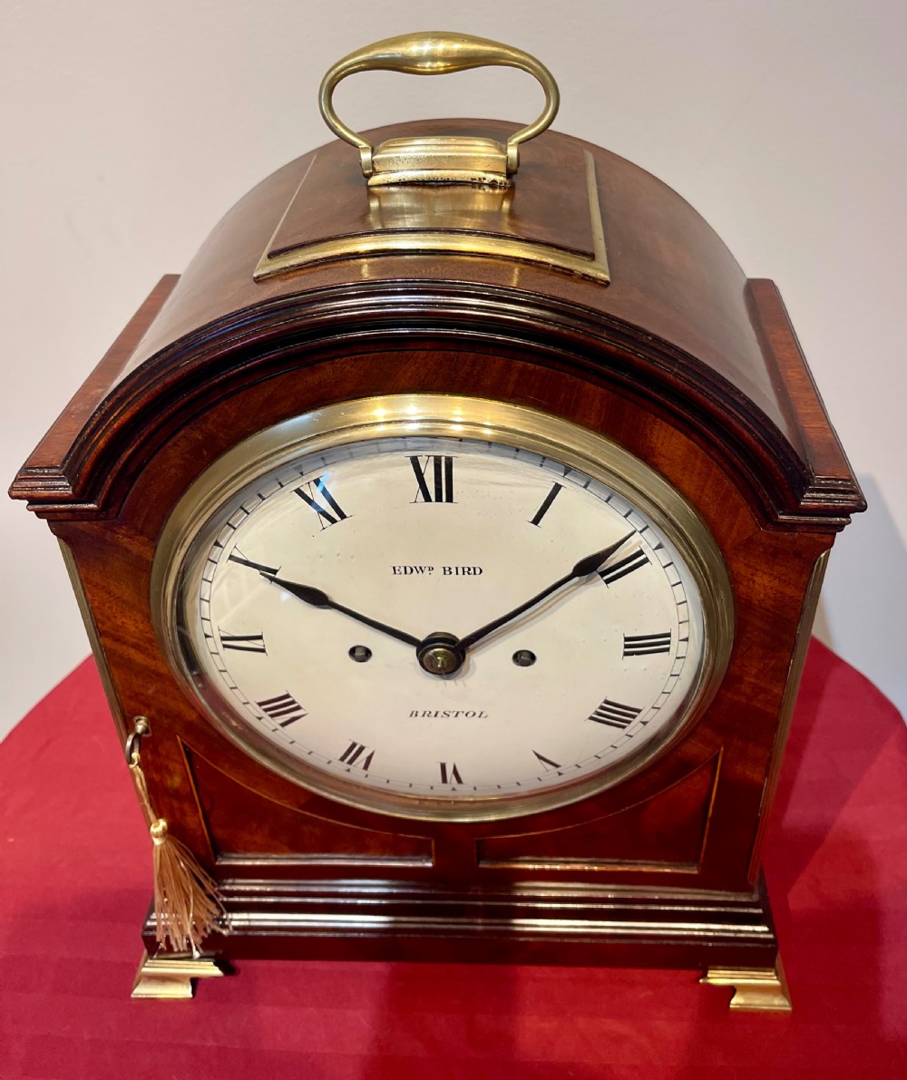 A George III mahogany bracket clock featuring an arched top with a brass handle, a circular white dial with Roman numerals, and brass bracket feet. View V
