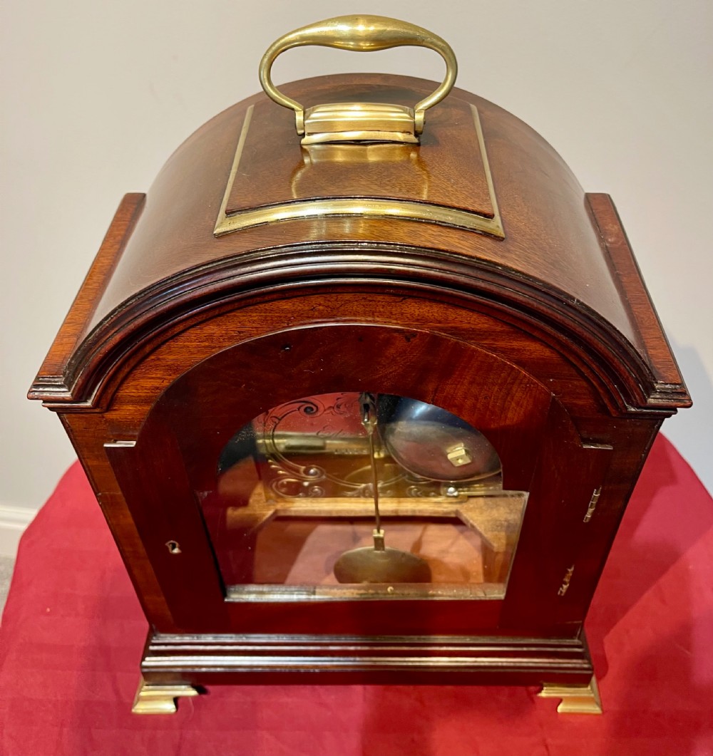 A George III mahogany bracket clock featuring an arched top with a brass handle, a circular white dial with Roman numerals, and brass bracket feet. View XII