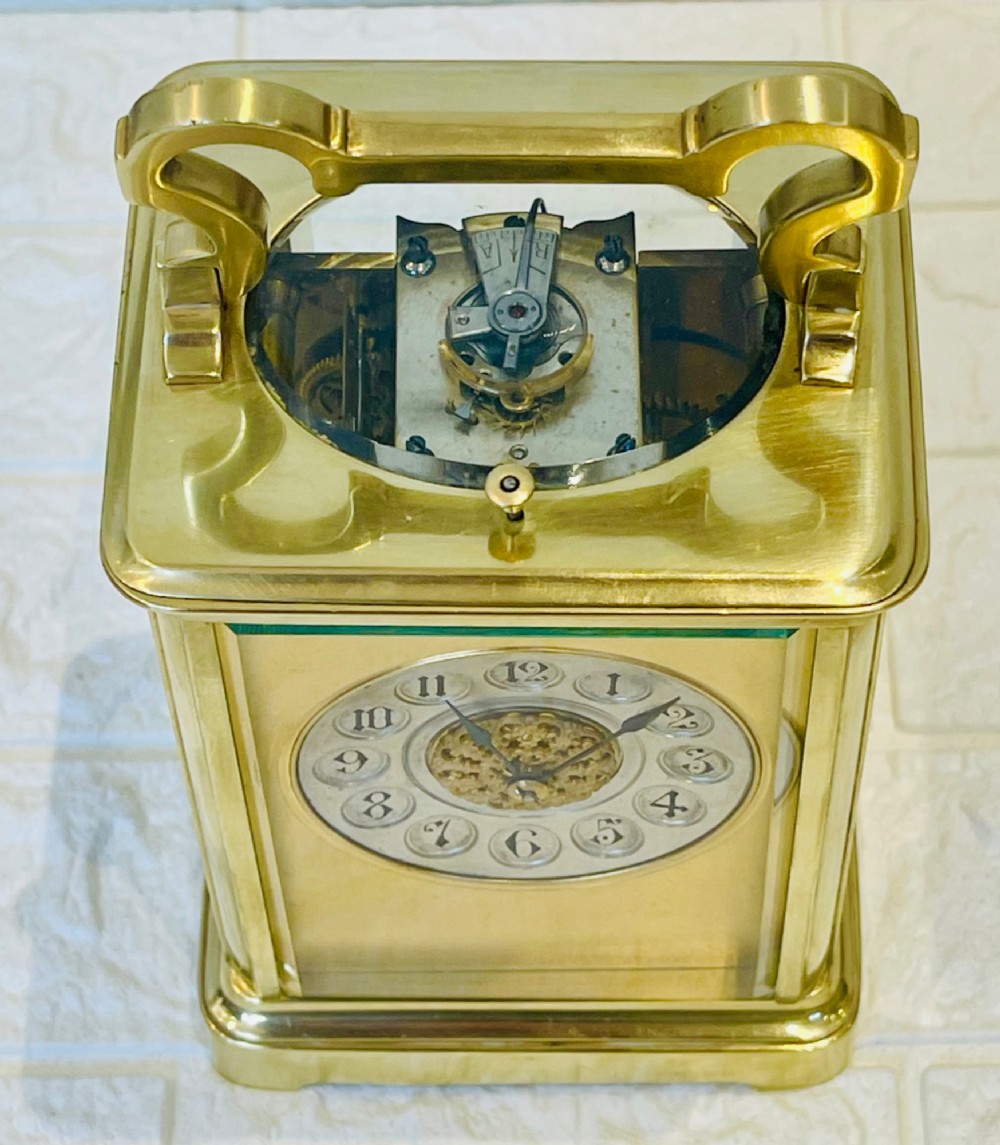 A polished brass repeating carriage clock featuring clear glass side panels, a scrolled top handle, and a silvered circular dial with Arabic numerals and a pierced gilt centre. View IV