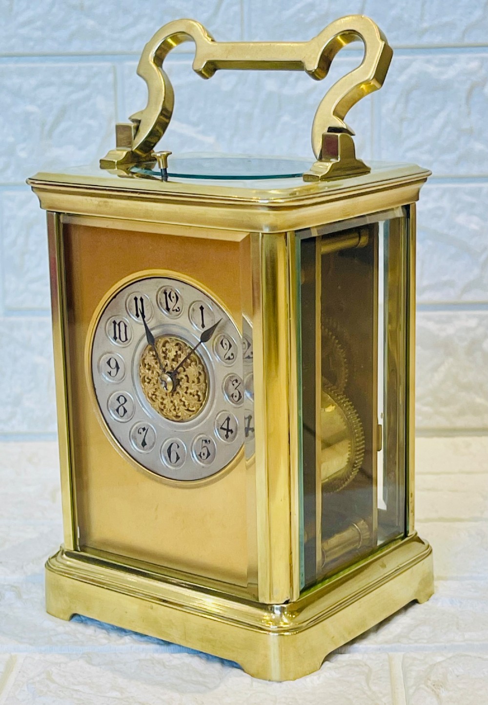 A polished brass repeating carriage clock featuring clear glass side panels, a scrolled top handle, and a silvered circular dial with Arabic numerals and a pierced gilt centre. View V