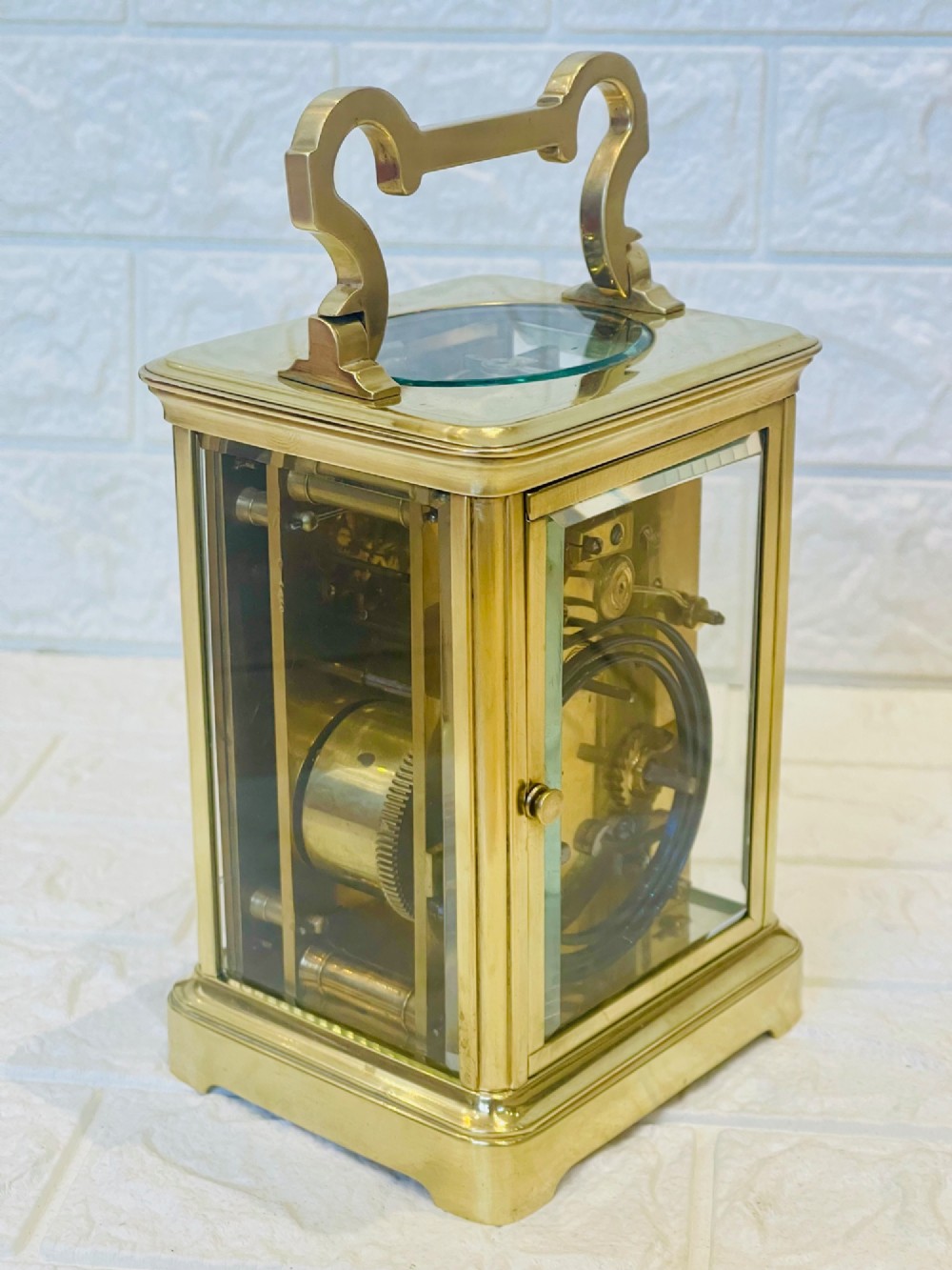 A polished brass repeating carriage clock featuring clear glass side panels, a scrolled top handle, and a silvered circular dial with Arabic numerals and a pierced gilt centre. View VII