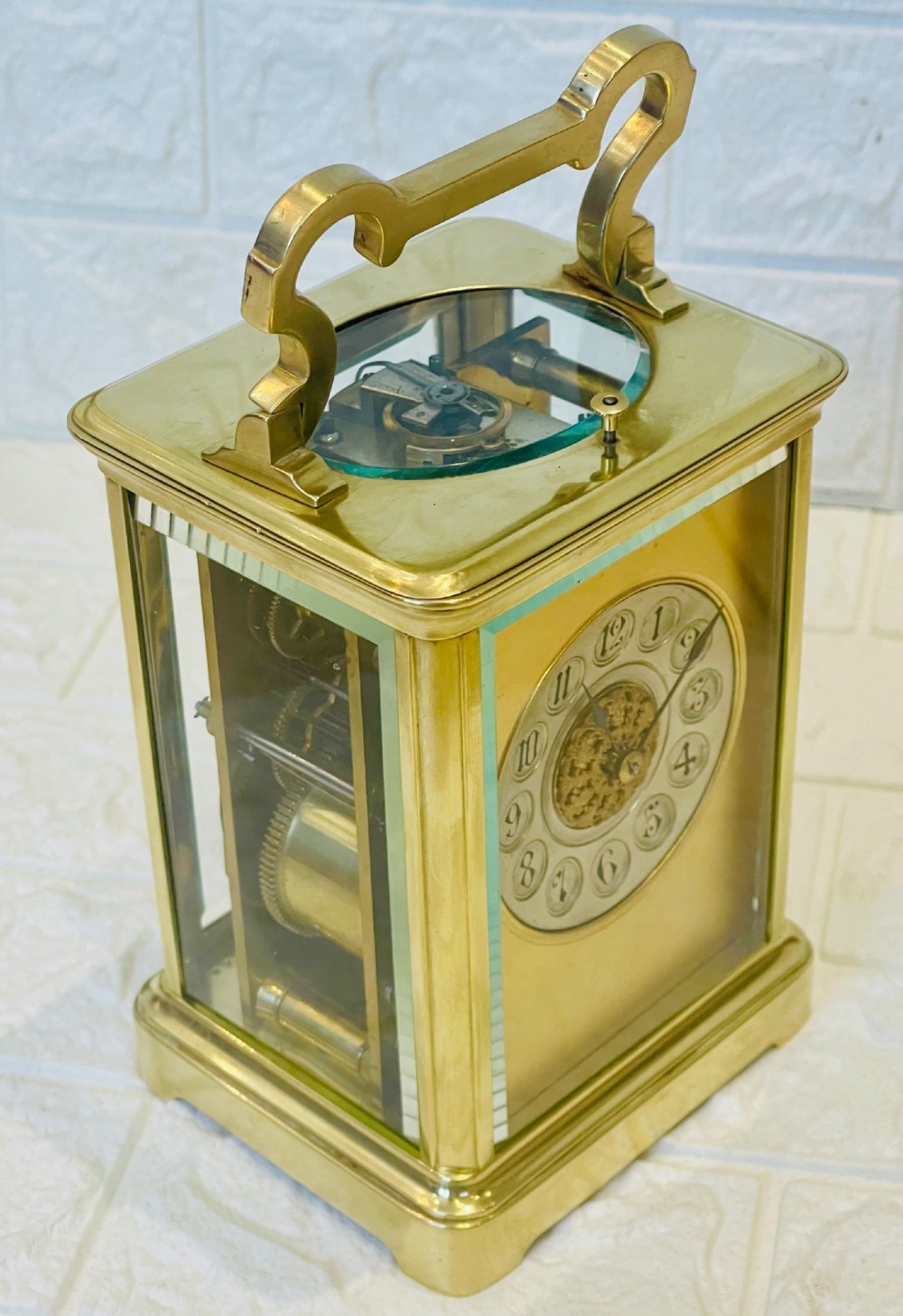 A polished brass repeating carriage clock featuring clear glass side panels, a scrolled top handle, and a silvered circular dial with Arabic numerals and a pierced gilt centre. View XII