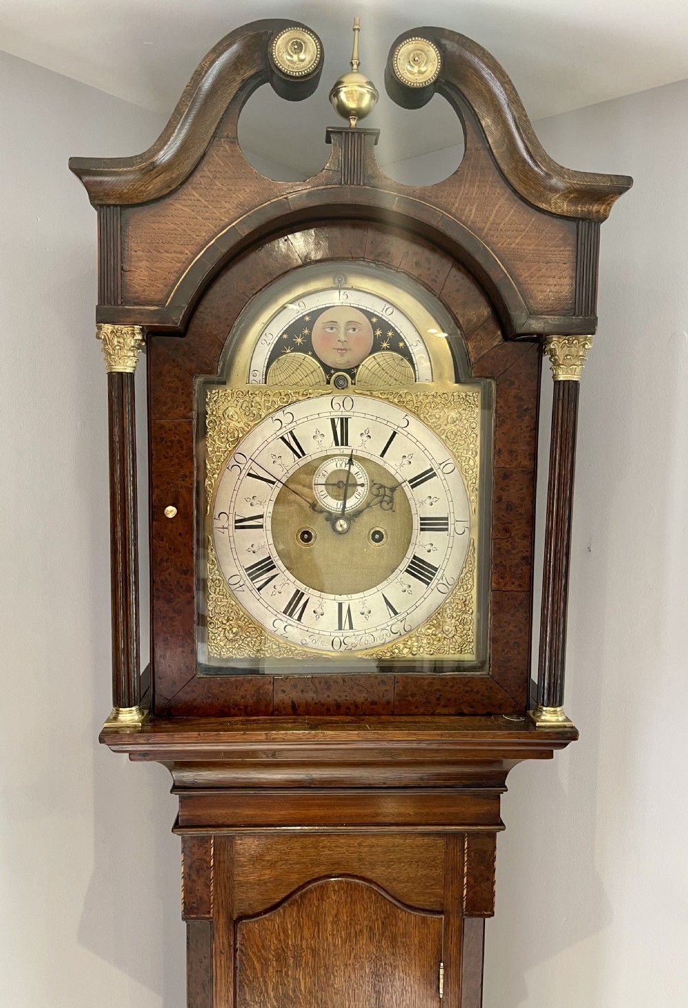 An antique Georgian longcase clock featuring a walnut and oak case, swan-neck pediment, brass finial, and an engraved brass dial with a moonphase display. View V