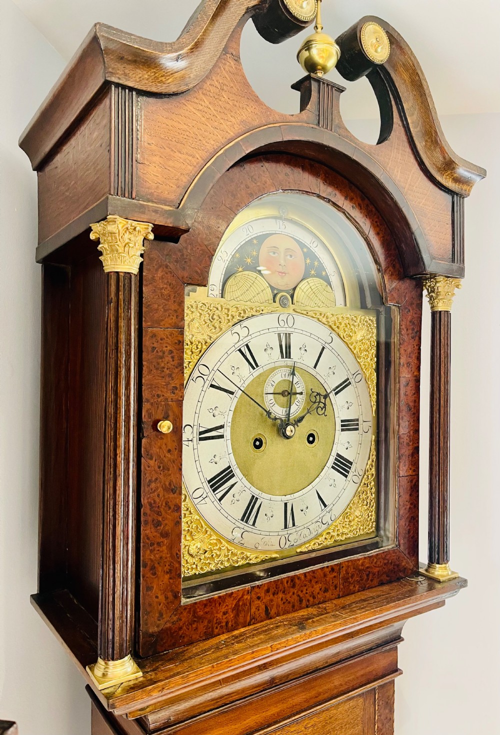 An antique Georgian longcase clock featuring a walnut and oak case, swan-neck pediment, brass finial, and an engraved brass dial with a moonphase display. View VI