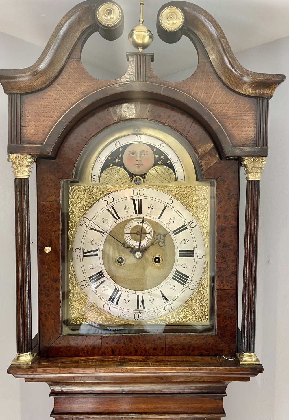 An antique Georgian longcase clock featuring a walnut and oak case, swan-neck pediment, brass finial, and an engraved brass dial with a moonphase display. View VII