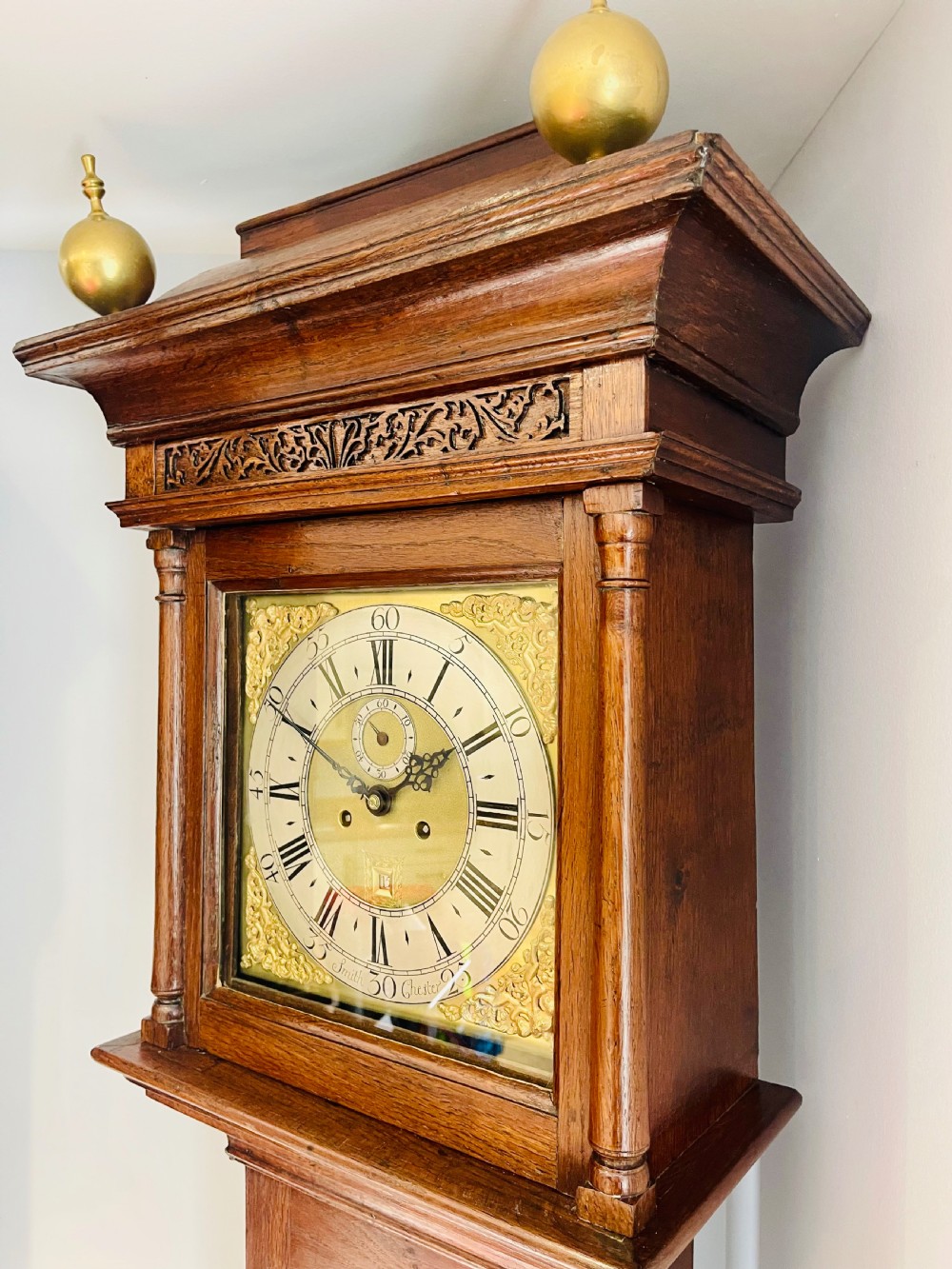 An 18th-century Georgian oak longcase clock featuring a square brass dial with silvered chapter ring, brass spandrels, twin brass finials, and a tall slender timber case. View V