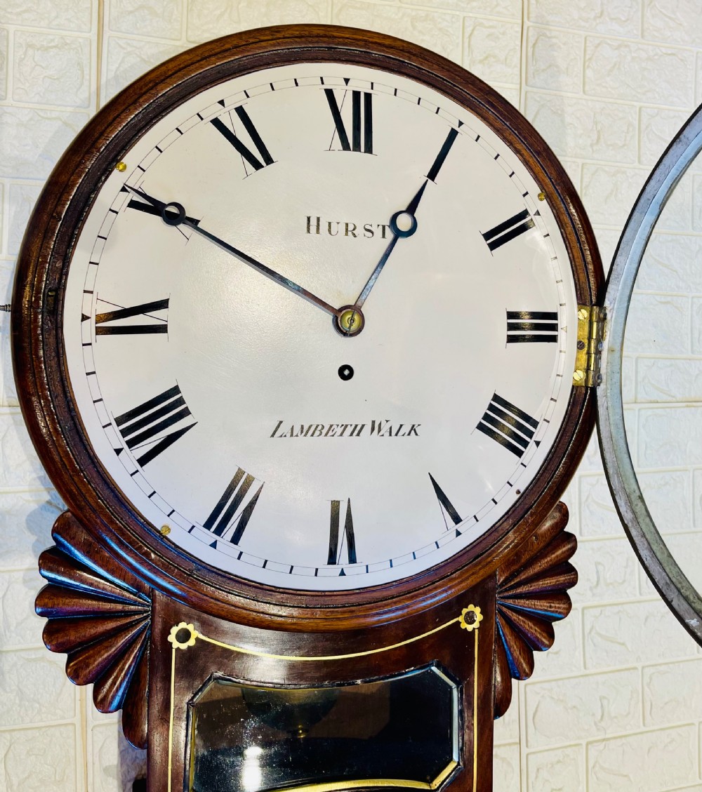 An antique Regency mahogany drop dial wall clock featuring brass line inlay, carved fan side ears, and a cream dial with Roman numerals. View XIV