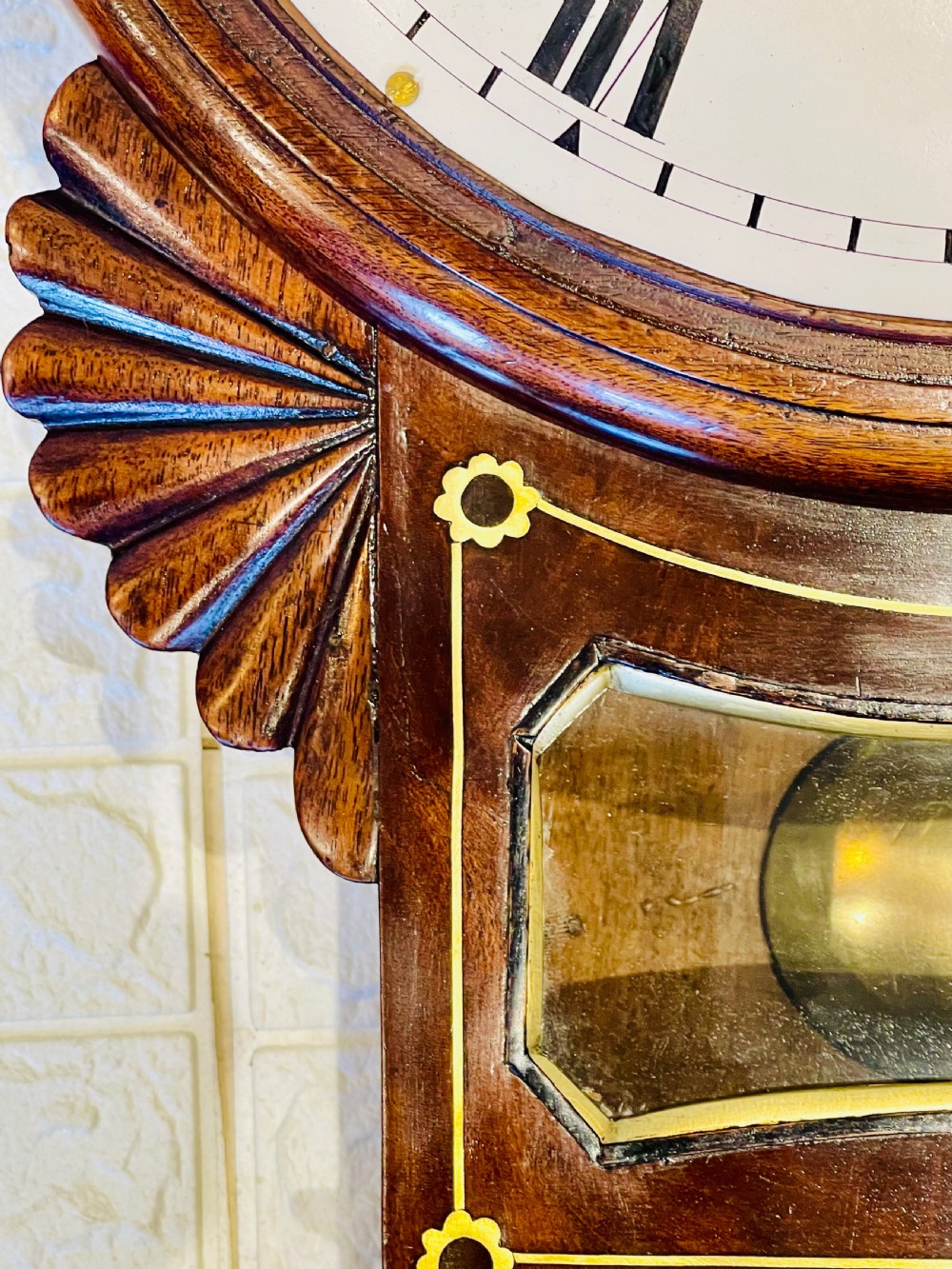 An antique Regency mahogany drop dial wall clock featuring brass line inlay, carved fan side ears, and a cream dial with Roman numerals. View XV