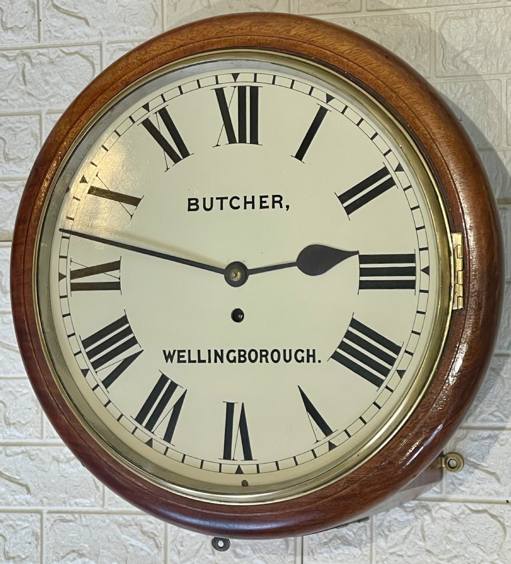 A circular Victorian oak wall clock featuring a cream painted dial with Roman numerals, black hands, a brass bezel, and the maker inscription Butcher, Wellingborough.