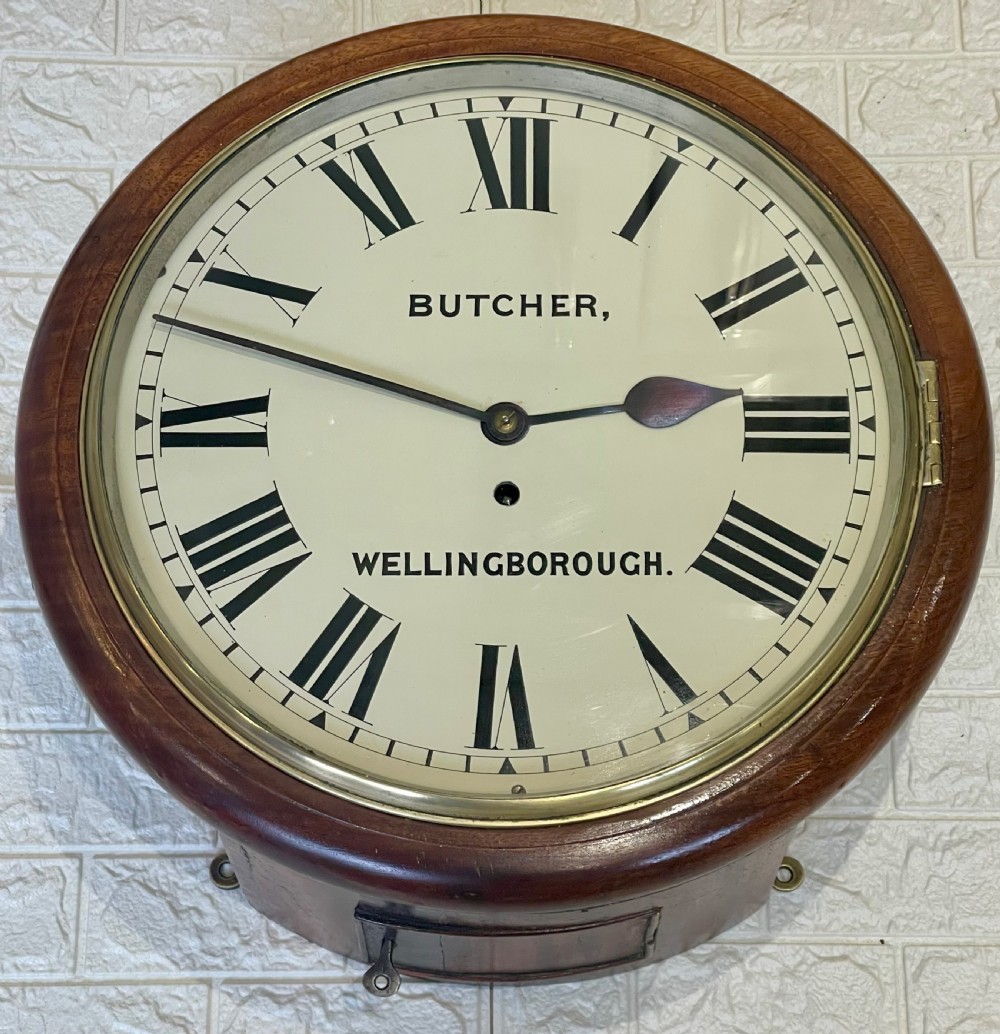 A circular Victorian oak wall clock featuring a cream painted dial with Roman numerals, black hands, a brass bezel, and the maker inscription Butcher, Wellingborough. View II