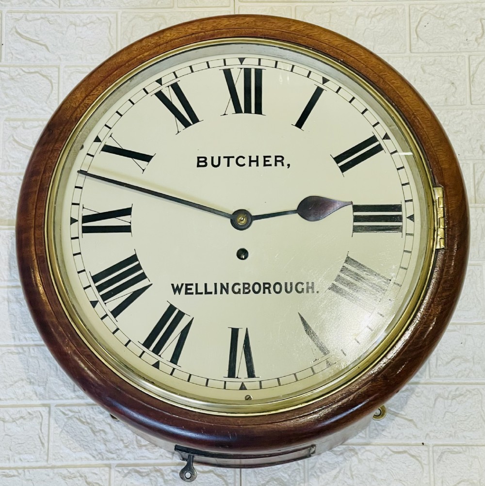 A circular Victorian oak wall clock featuring a cream painted dial with Roman numerals, black hands, a brass bezel, and the maker inscription Butcher, Wellingborough. View III