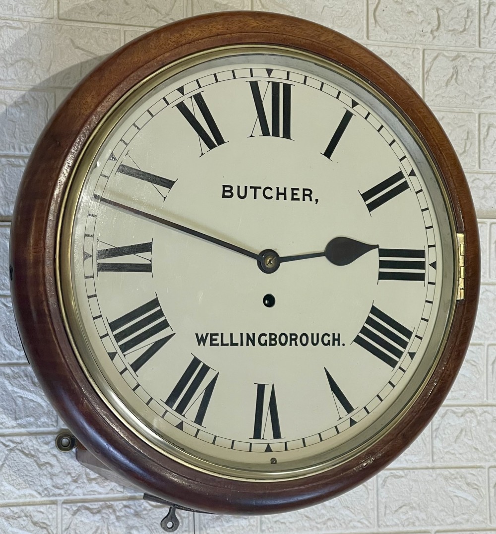 A circular Victorian oak wall clock featuring a cream painted dial with Roman numerals, black hands, a brass bezel, and the maker inscription Butcher, Wellingborough. View IV