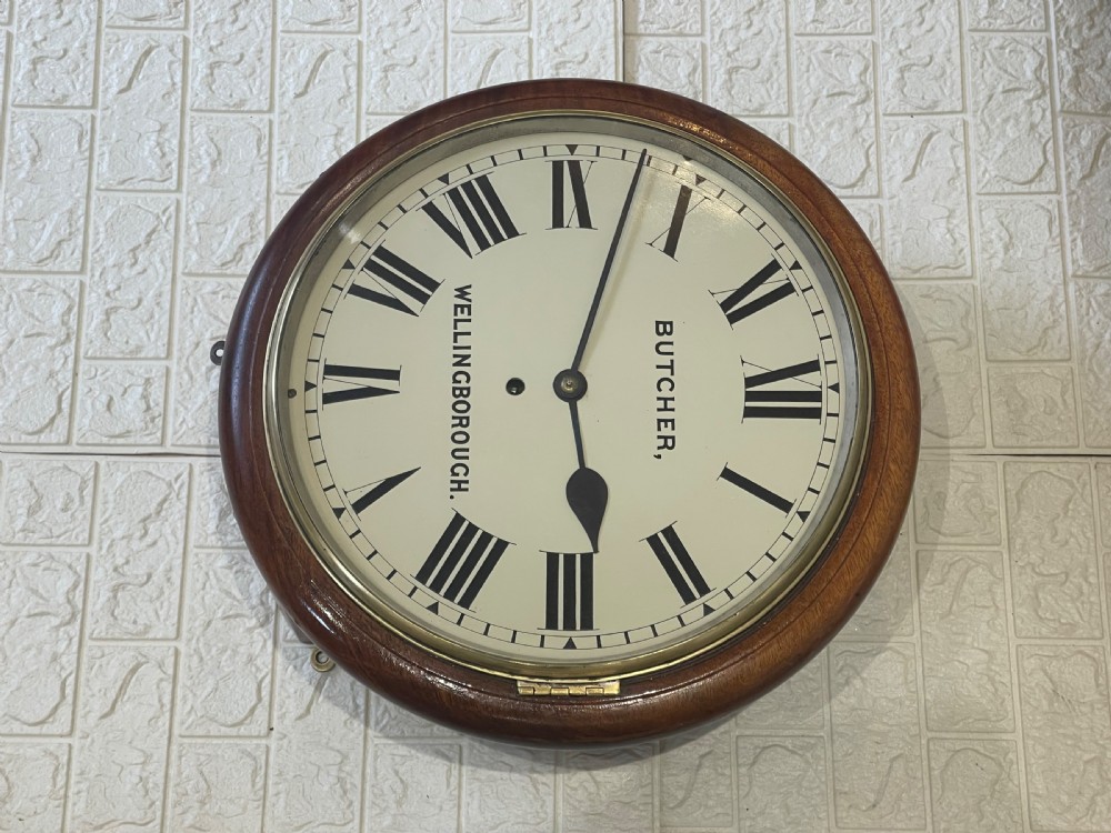 A circular Victorian oak wall clock featuring a cream painted dial with Roman numerals, black hands, a brass bezel, and the maker inscription Butcher, Wellingborough. View V