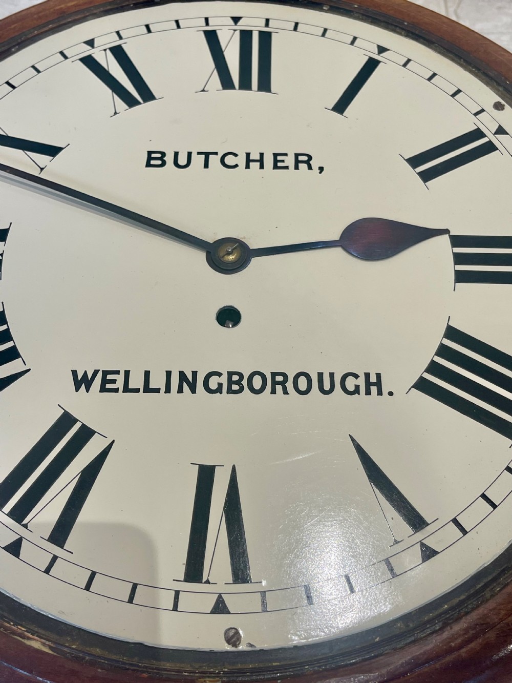 A circular Victorian oak wall clock featuring a cream painted dial with Roman numerals, black hands, a brass bezel, and the maker inscription Butcher, Wellingborough. View X