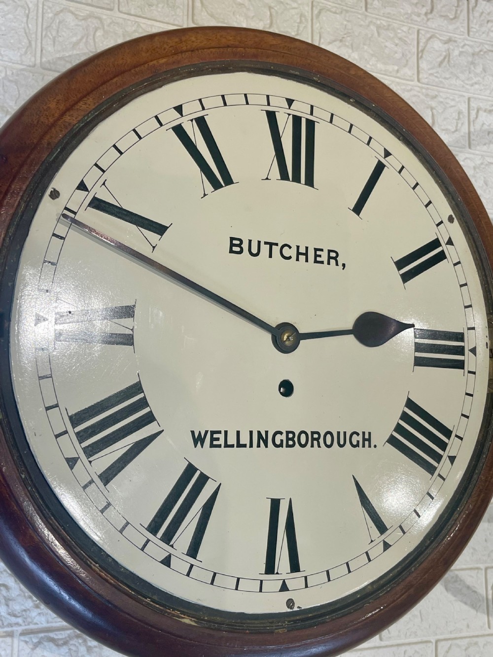 A circular Victorian oak wall clock featuring a cream painted dial with Roman numerals, black hands, a brass bezel, and the maker inscription Butcher, Wellingborough. View XI