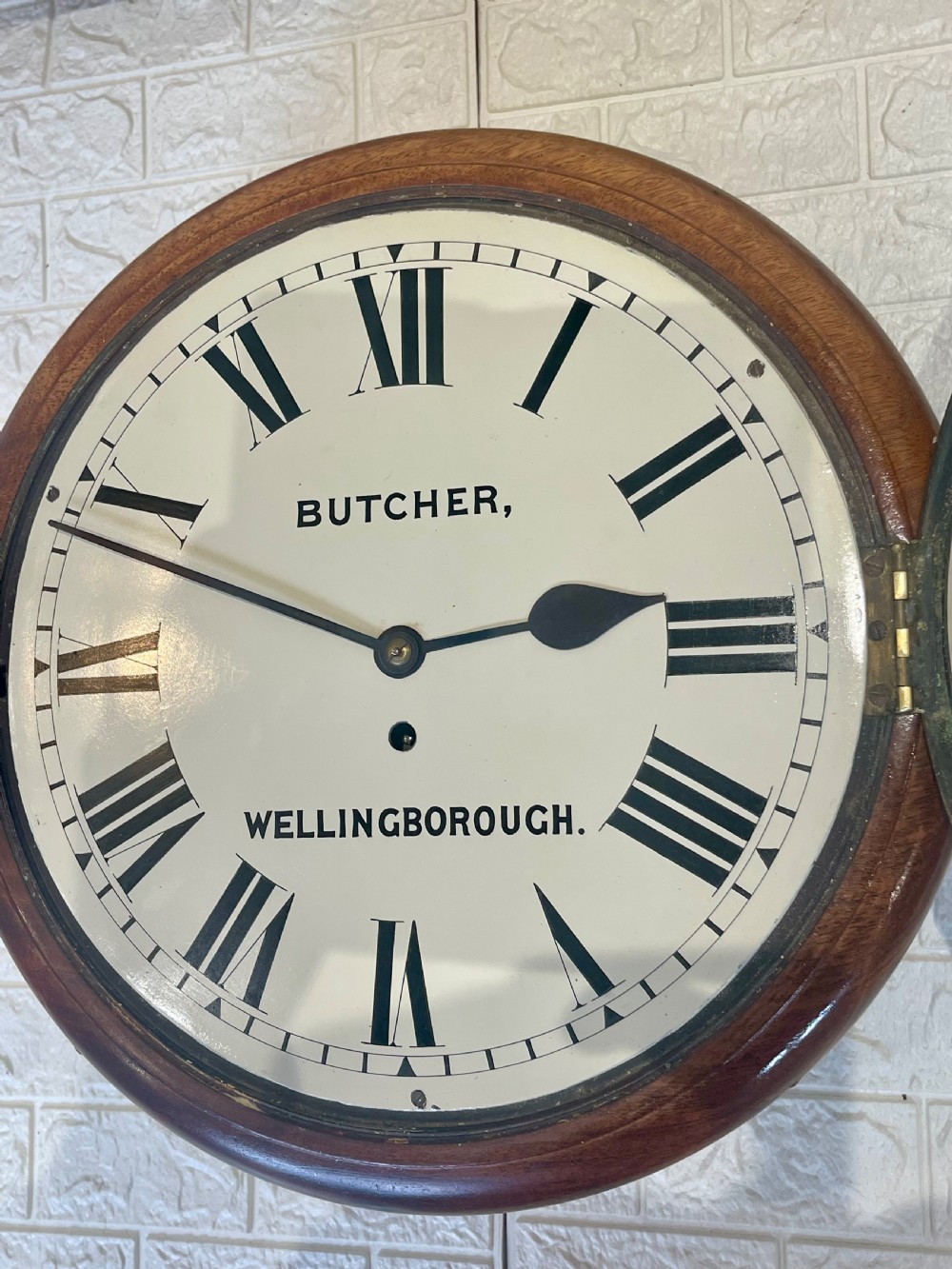 A circular Victorian oak wall clock featuring a cream painted dial with Roman numerals, black hands, a brass bezel, and the maker inscription Butcher, Wellingborough. View XII