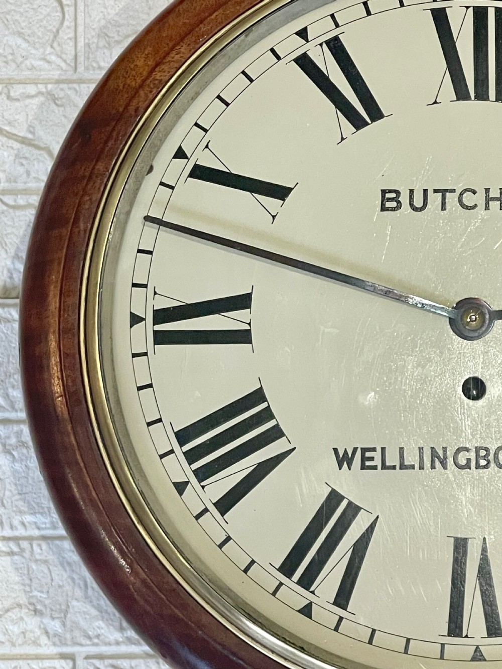 A circular Victorian oak wall clock featuring a cream painted dial with Roman numerals, black hands, a brass bezel, and the maker inscription Butcher, Wellingborough. View XIII