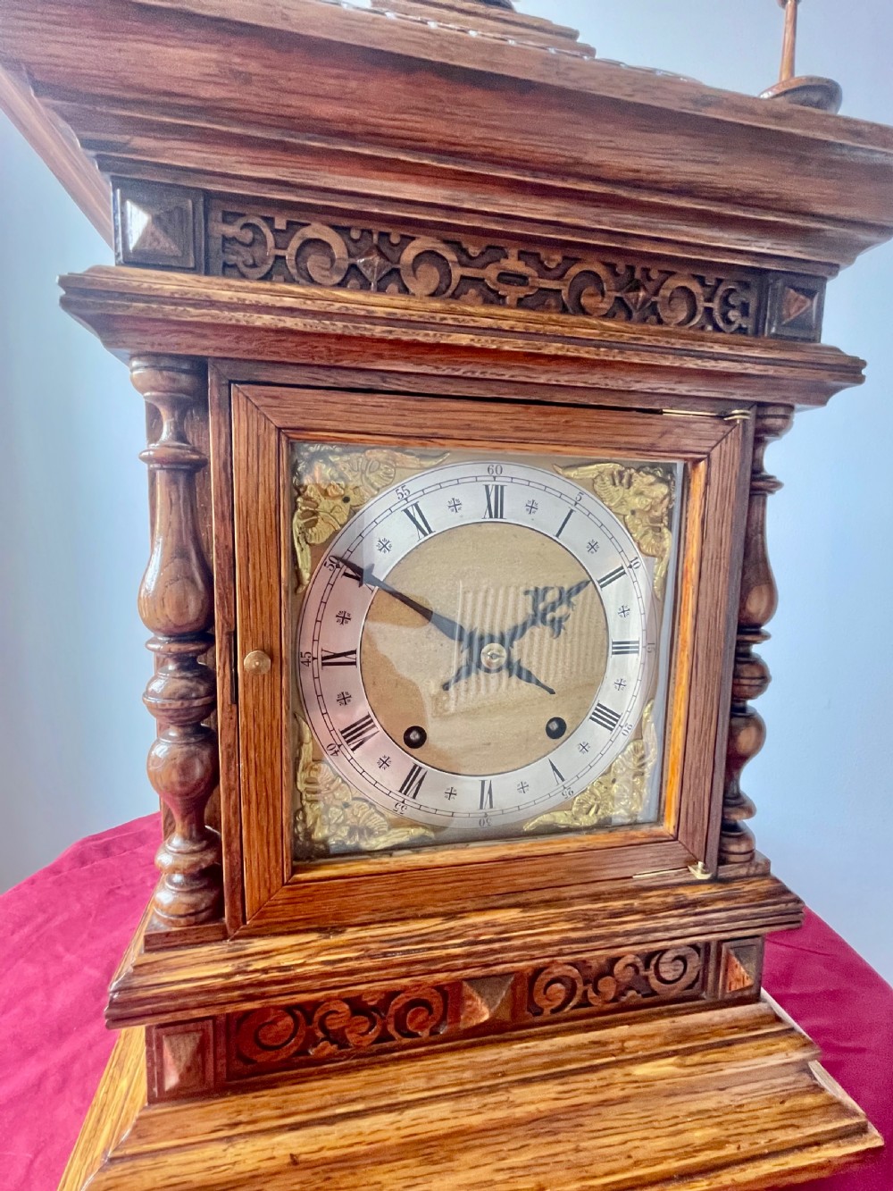 An antique carved oak bracket clock featuring finials, turned columns, and a brass dial with silvered chapter ring. View V