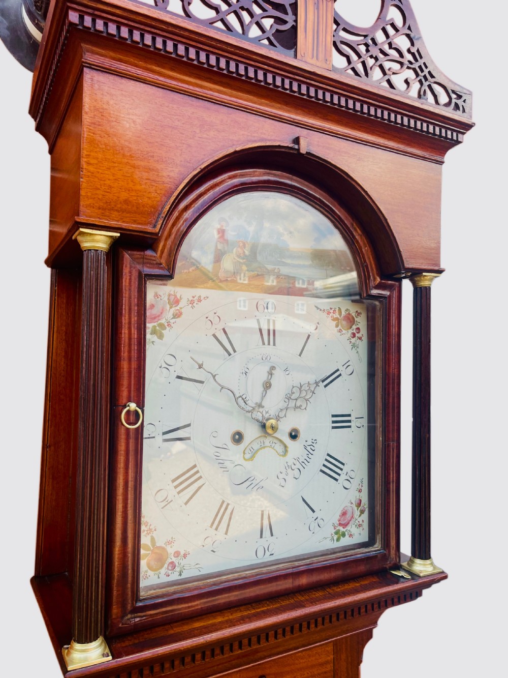 A Georgian oak longcase clock featuring a fretwork pediment, brass finial, hand-painted dial with Roman numerals, and an arched waist door. View IV