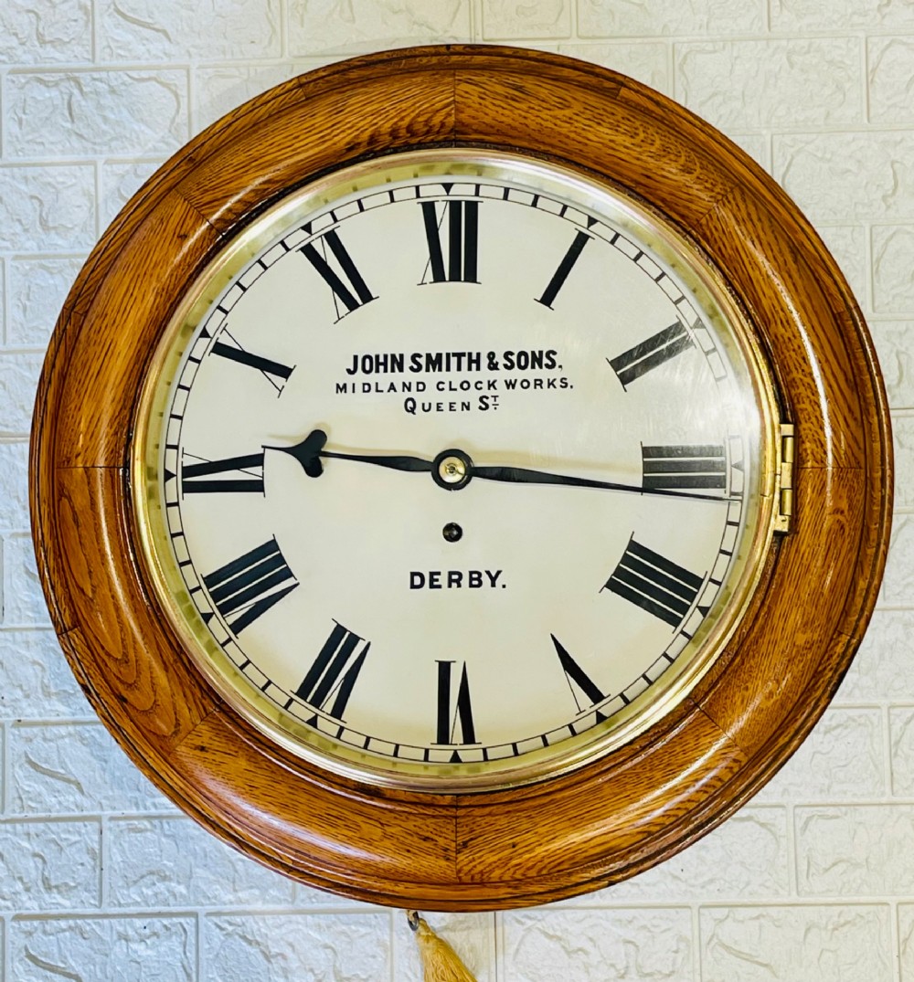 A circular antique oak wall clock featuring a white painted dial with black Roman numerals, brass bezel, and maker inscription.