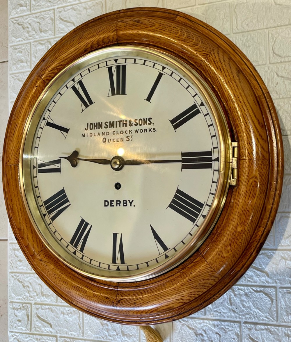 A circular antique oak wall clock featuring a white painted dial with black Roman numerals, brass bezel, and maker inscription. View II