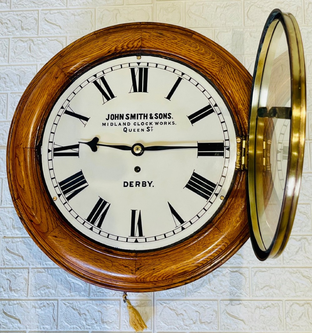 A circular antique oak wall clock featuring a white painted dial with black Roman numerals, brass bezel, and maker inscription. View IV
