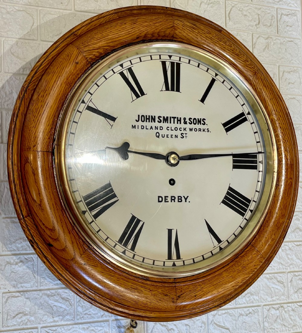 A circular antique oak wall clock featuring a white painted dial with black Roman numerals, brass bezel, and maker inscription. View V