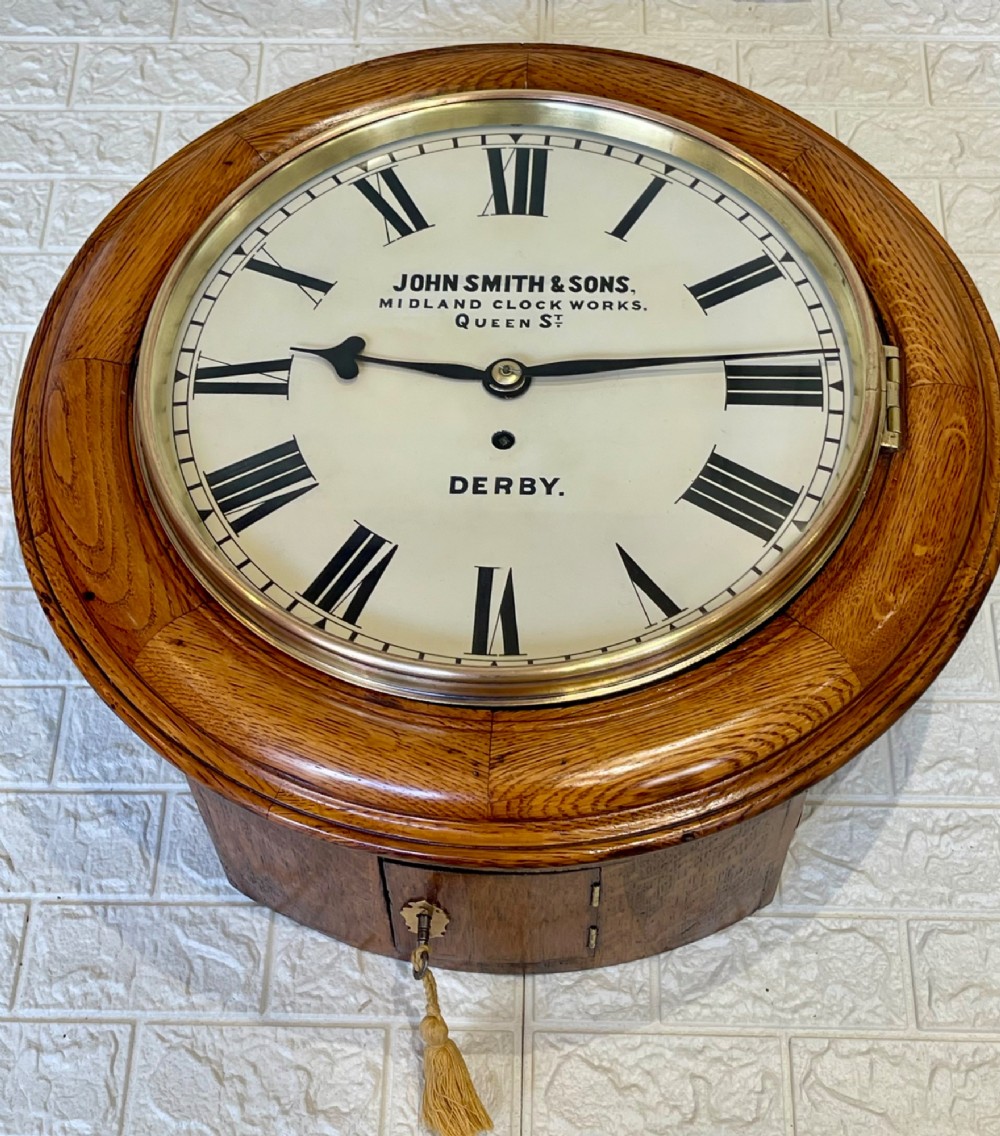 A circular antique oak wall clock featuring a white painted dial with black Roman numerals, brass bezel, and maker inscription. View VI