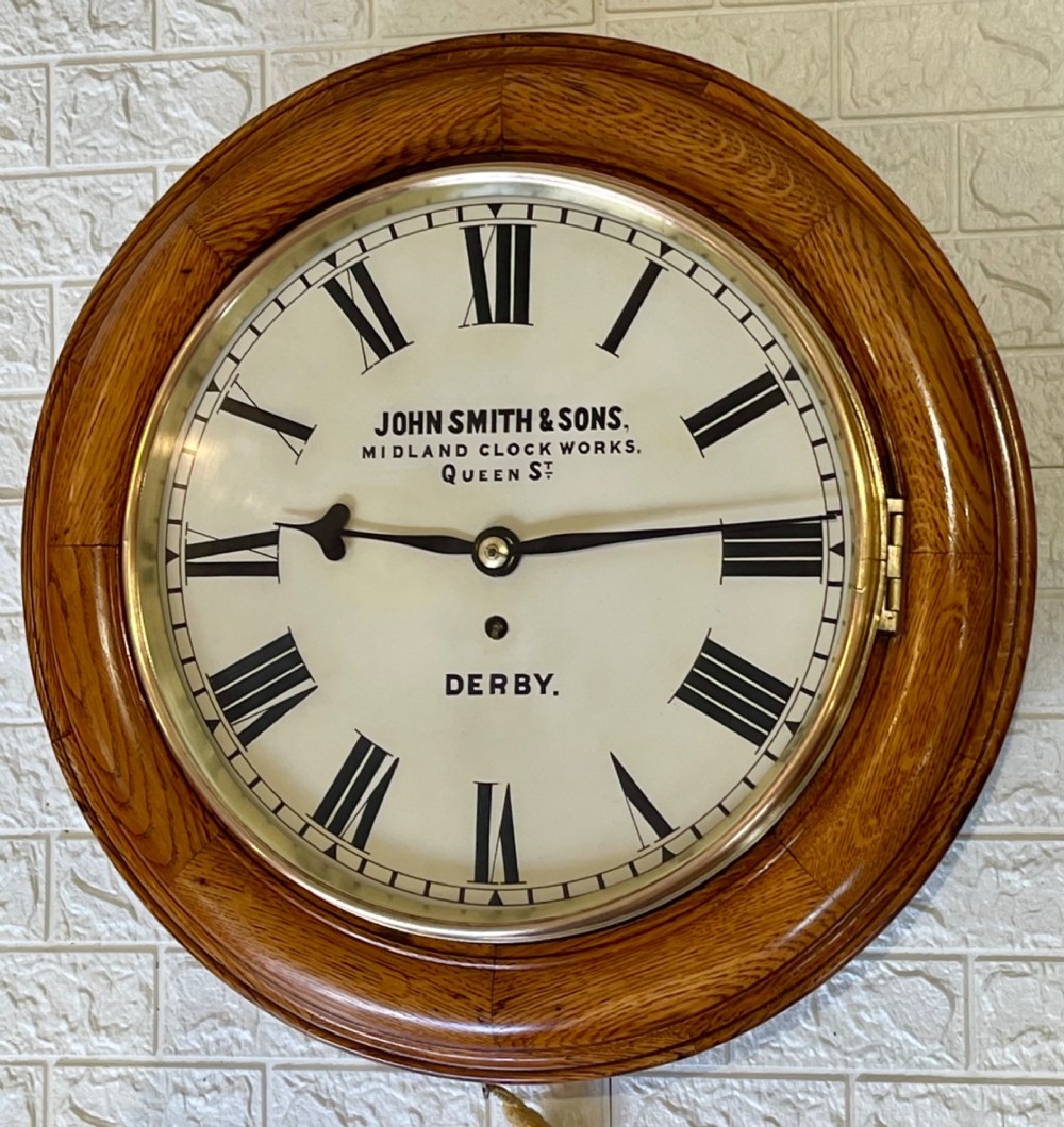 A circular antique oak wall clock featuring a white painted dial with black Roman numerals, brass bezel, and maker inscription. View VII