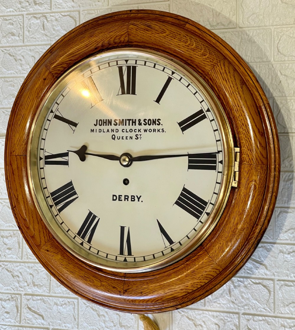 A circular antique oak wall clock featuring a white painted dial with black Roman numerals, brass bezel, and maker inscription. View VIII