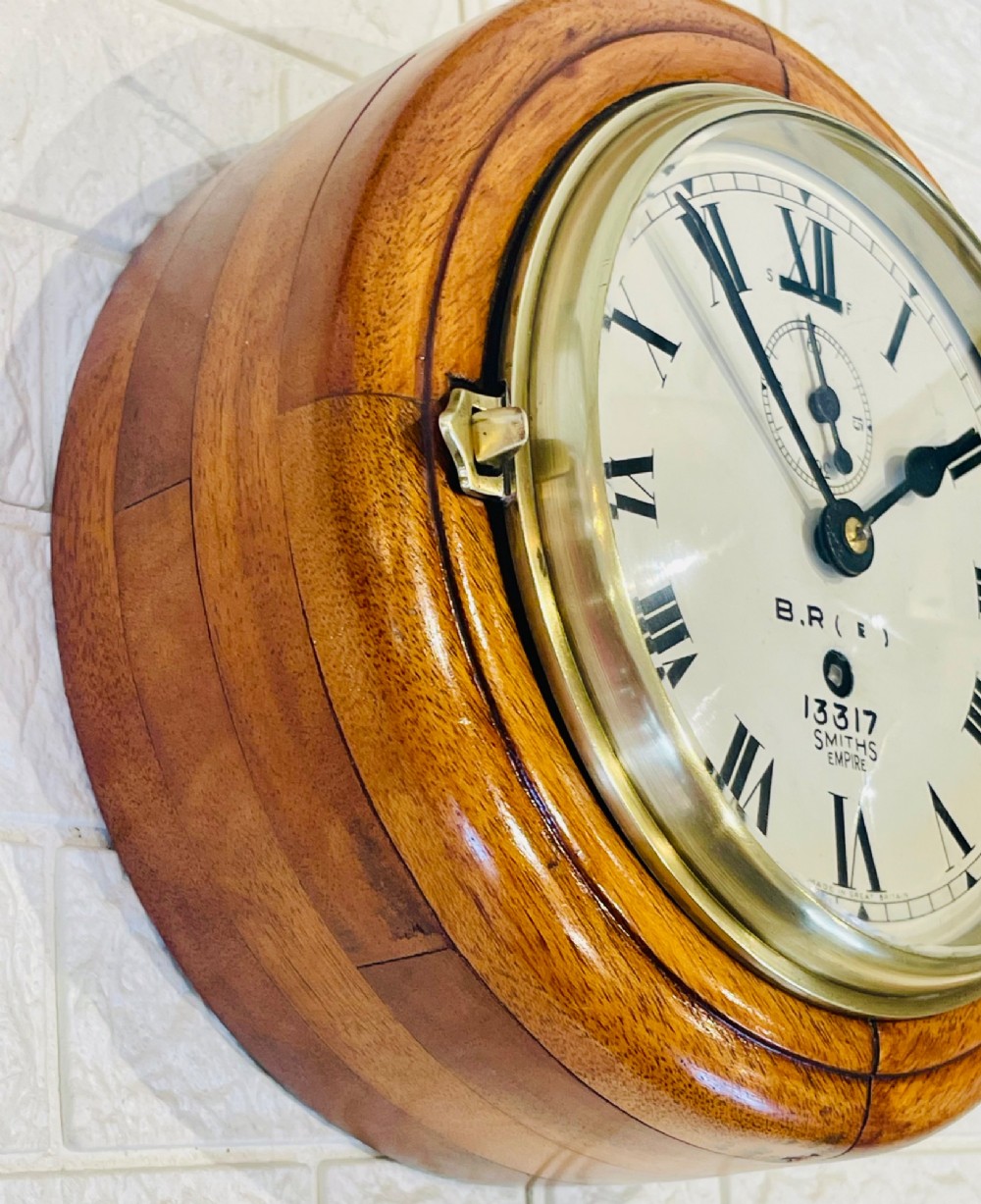 A circular vintage British Rail wall clock featuring a warm brown wooden casing, a hinged brass bezel, and a white dial with black Roman numerals stamped B.R.(E) Smiths Empire. View II