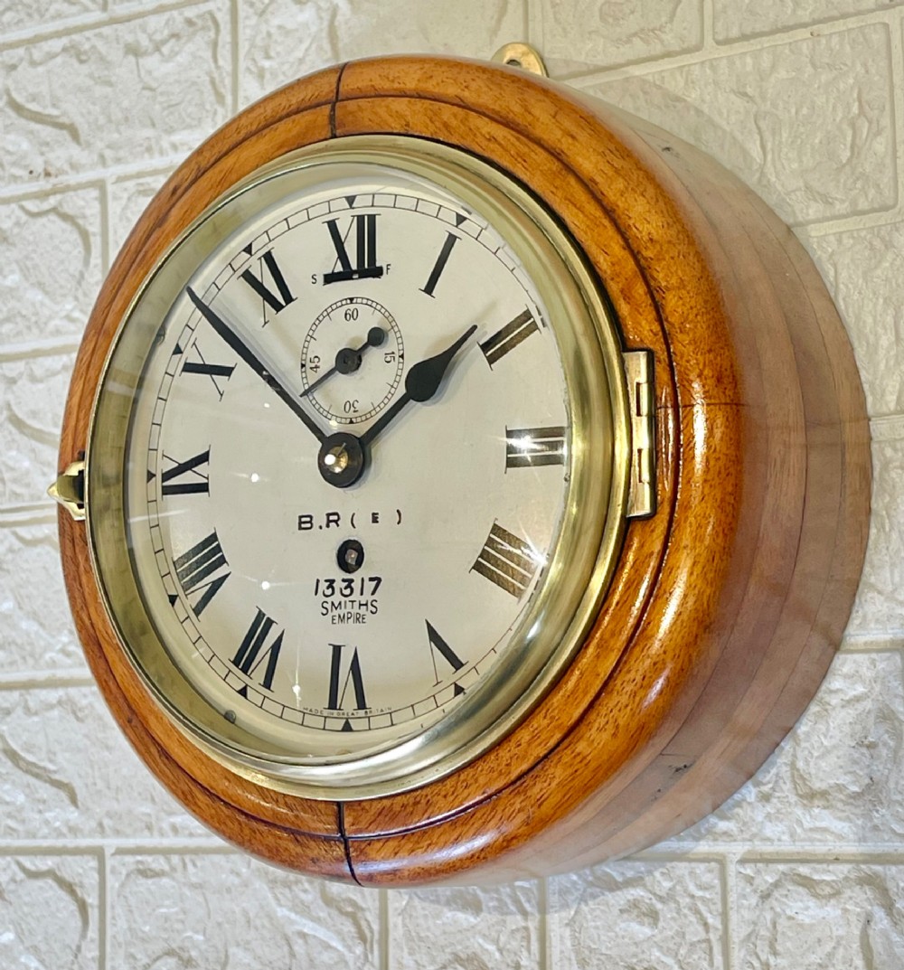 A circular vintage British Rail wall clock featuring a warm brown wooden casing, a hinged brass bezel, and a white dial with black Roman numerals stamped B.R.(E) Smiths Empire. View V