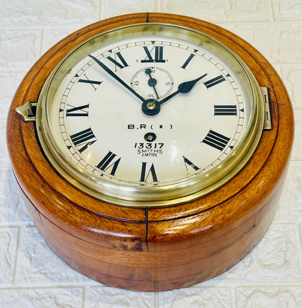 A circular vintage British Rail wall clock featuring a warm brown wooden casing, a hinged brass bezel, and a white dial with black Roman numerals stamped B.R.(E) Smiths Empire. View IX