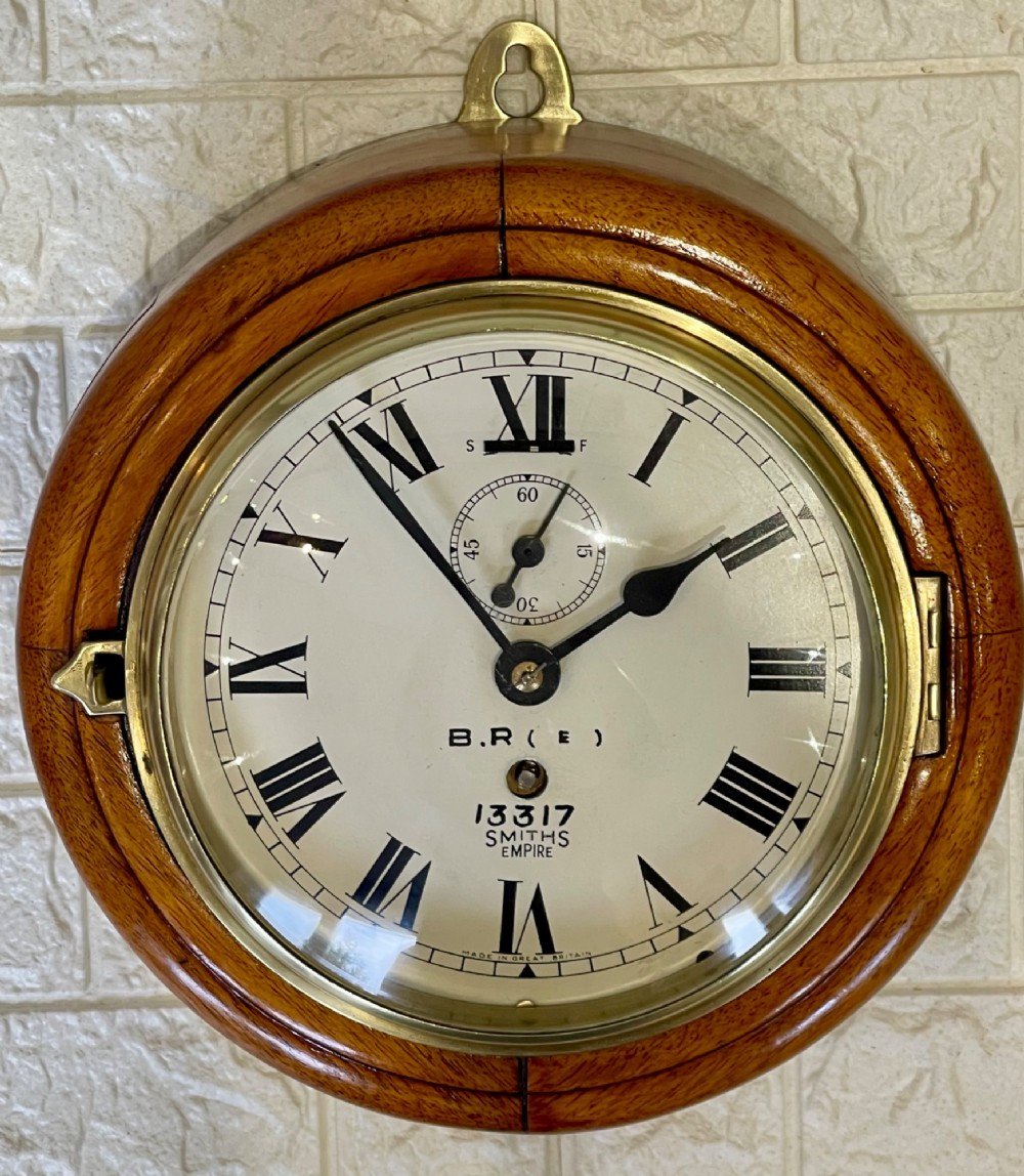 A circular vintage British Rail wall clock featuring a warm brown wooden casing, a hinged brass bezel, and a white dial with black Roman numerals stamped B.R.(E) Smiths Empire. View X