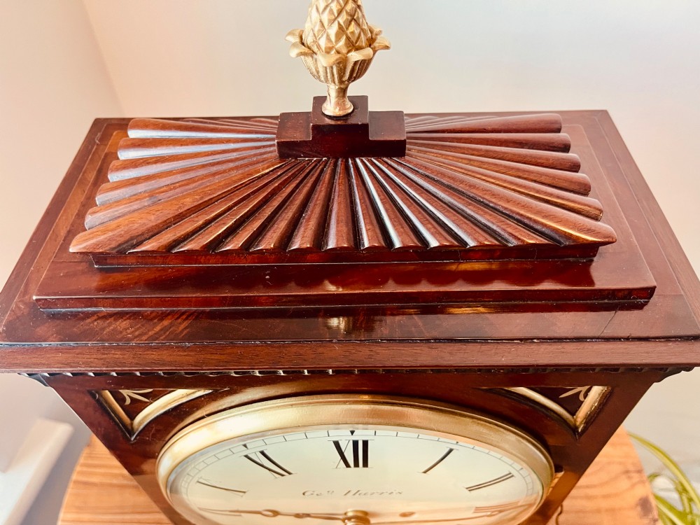 An antique Regency dark mahogany bracket clock featuring intricate brass inlay scrollwork, a brass pineapple finial, side brass lion-mask drop handles, and a cream dial with Roman numerals. View IV