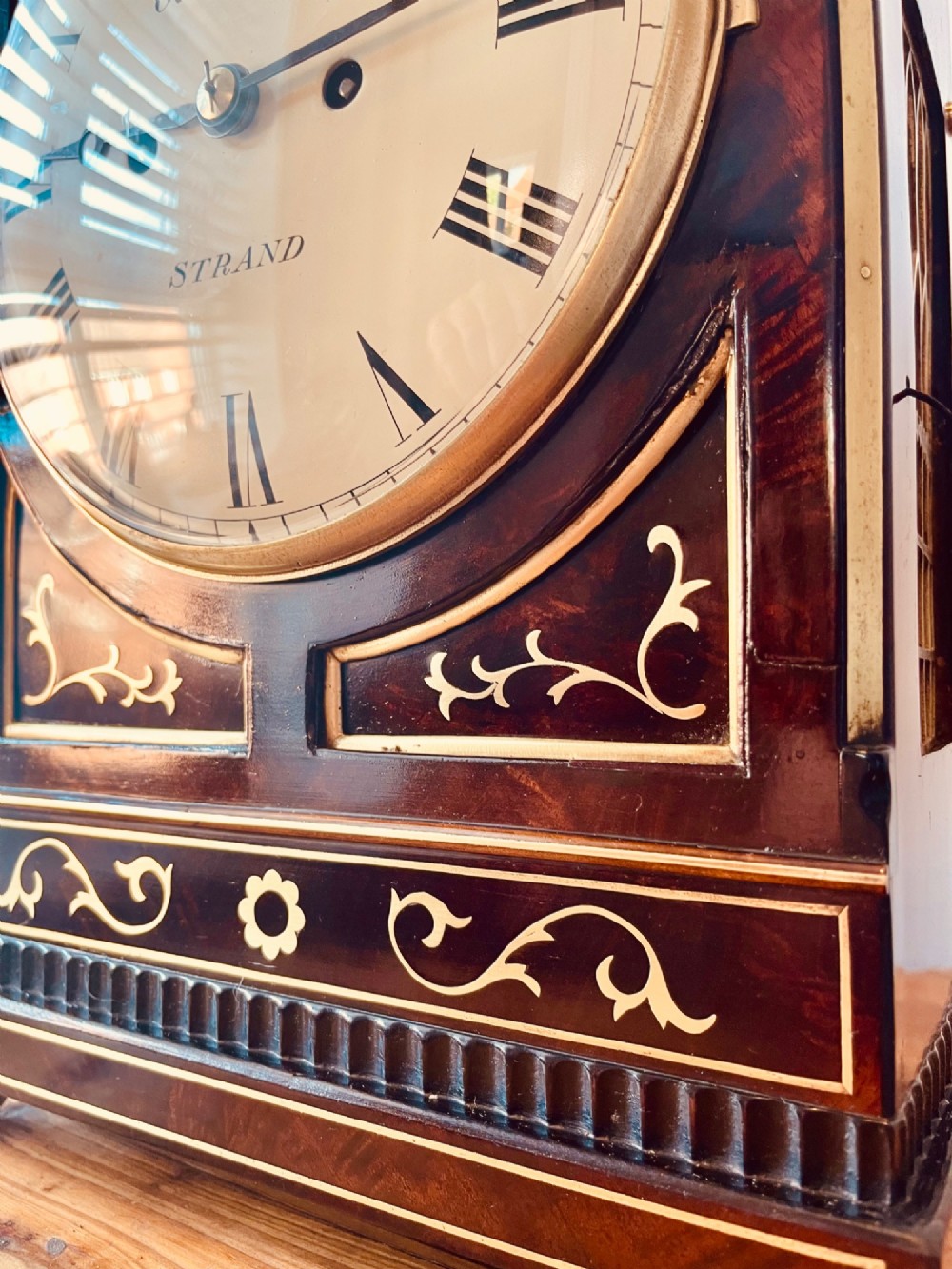 An antique Regency dark mahogany bracket clock featuring intricate brass inlay scrollwork, a brass pineapple finial, side brass lion-mask drop handles, and a cream dial with Roman numerals. View V