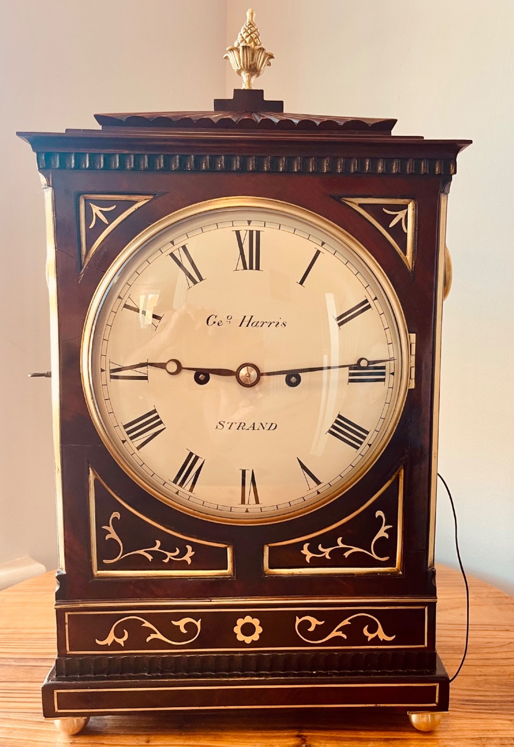 An antique Regency dark mahogany bracket clock featuring intricate brass inlay scrollwork, a brass pineapple finial, side brass lion-mask drop handles, and a cream dial with Roman numerals. View VII