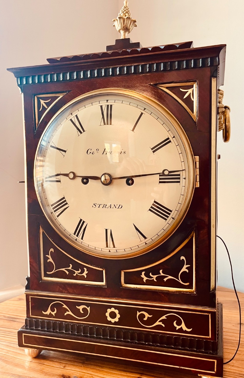 An antique Regency dark mahogany bracket clock featuring intricate brass inlay scrollwork, a brass pineapple finial, side brass lion-mask drop handles, and a cream dial with Roman numerals. View VIII