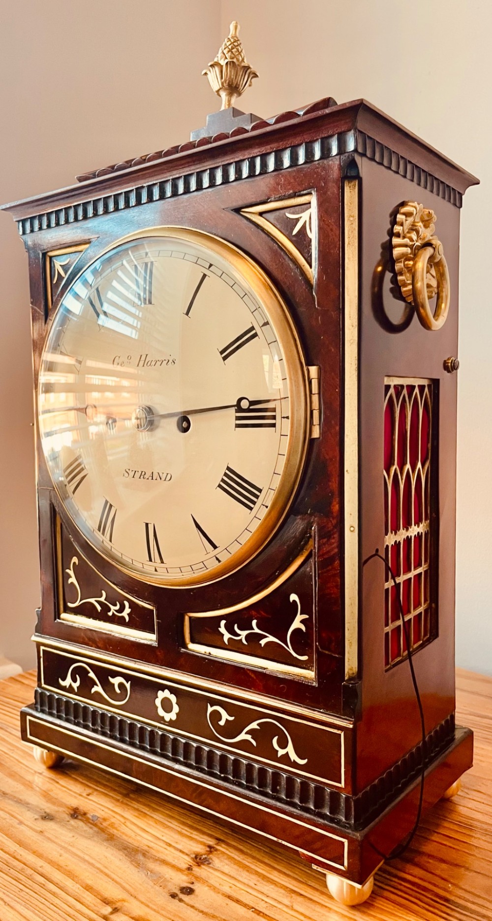 An antique Regency dark mahogany bracket clock featuring intricate brass inlay scrollwork, a brass pineapple finial, side brass lion-mask drop handles, and a cream dial with Roman numerals. View IX