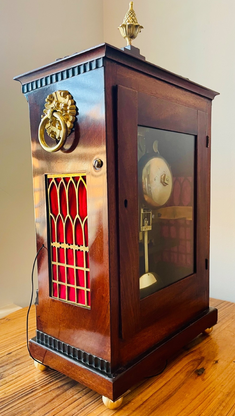 An antique Regency dark mahogany bracket clock featuring intricate brass inlay scrollwork, a brass pineapple finial, side brass lion-mask drop handles, and a cream dial with Roman numerals. View XI