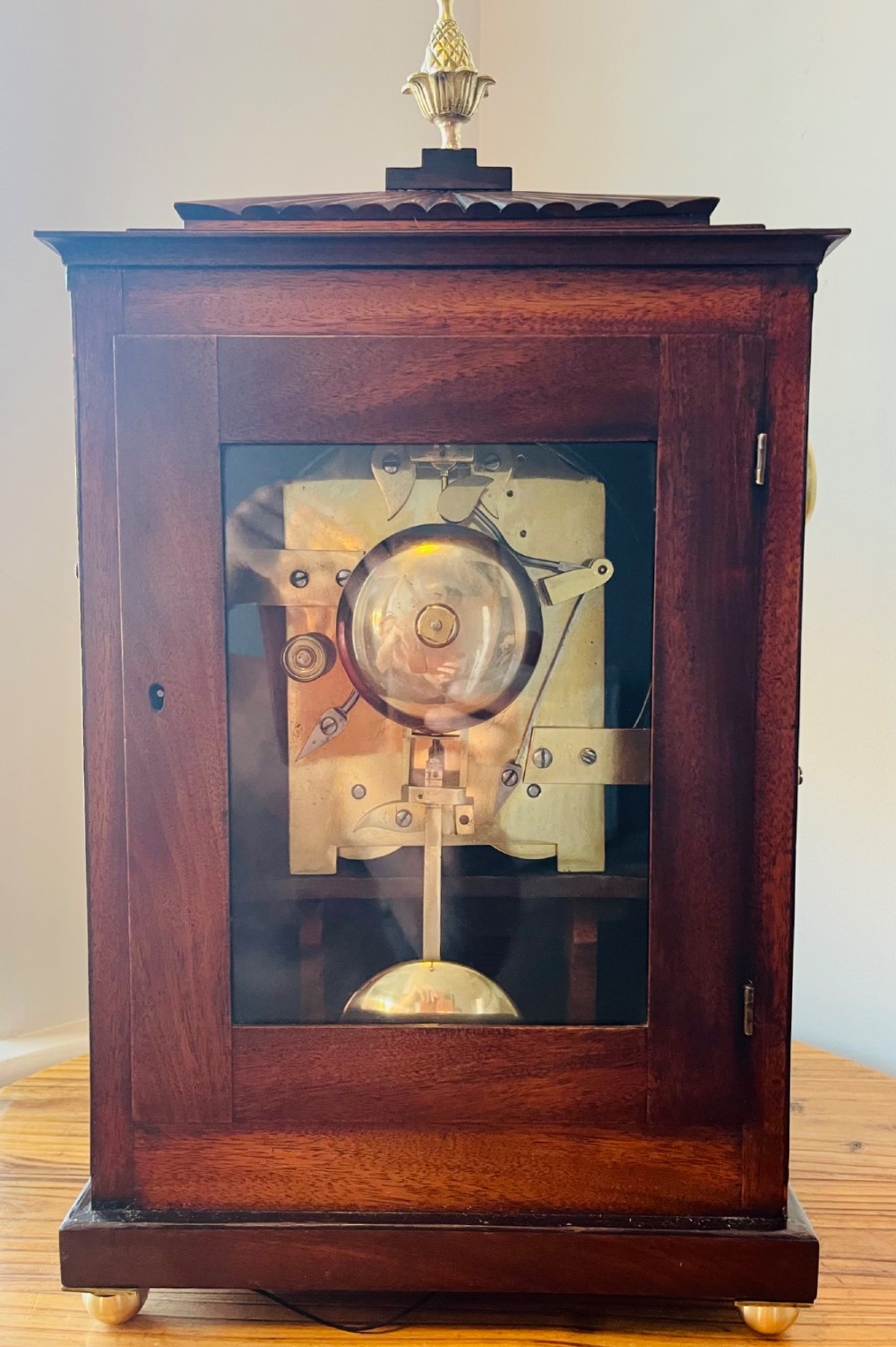 An antique Regency dark mahogany bracket clock featuring intricate brass inlay scrollwork, a brass pineapple finial, side brass lion-mask drop handles, and a cream dial with Roman numerals. View XII