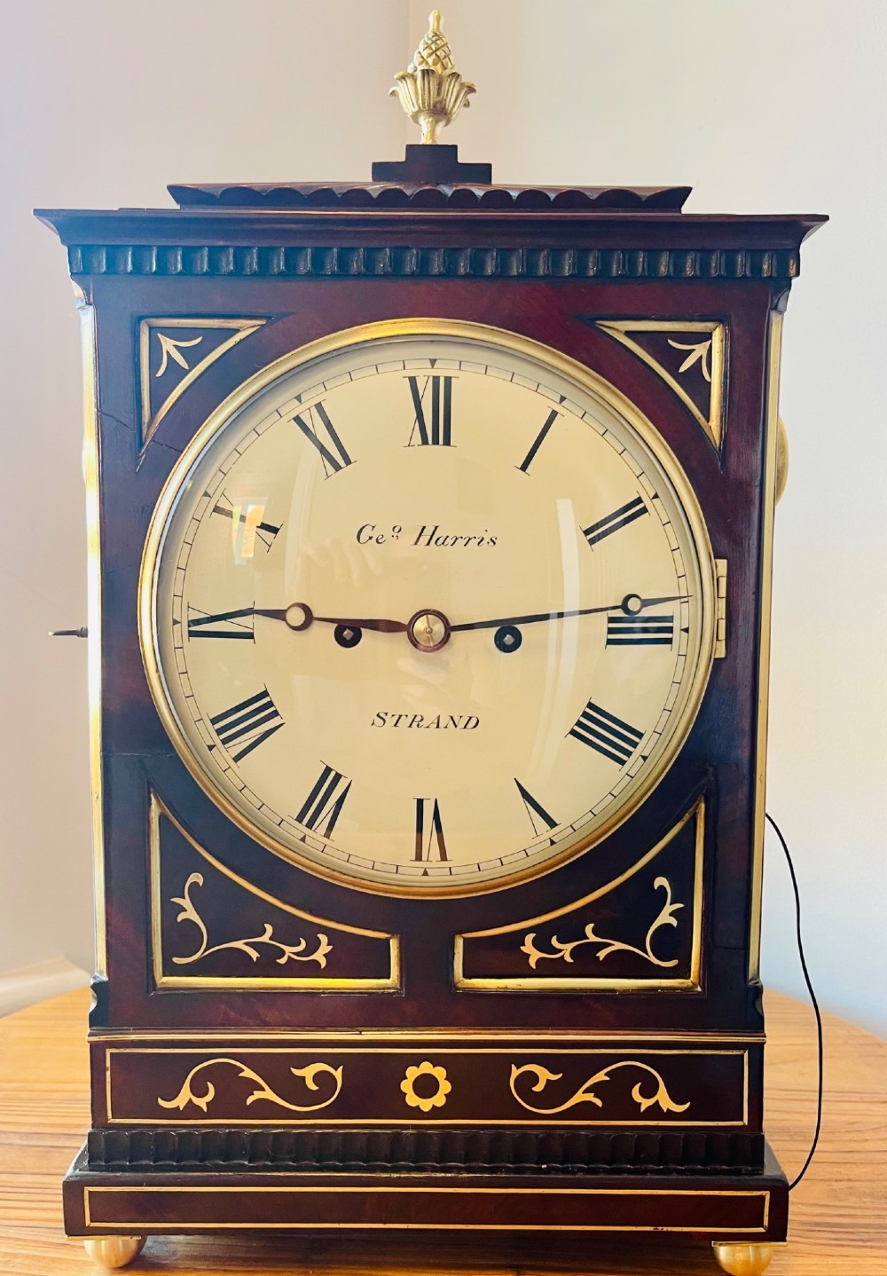 An antique Regency dark mahogany bracket clock featuring intricate brass inlay scrollwork, a brass pineapple finial, side brass lion-mask drop handles, and a cream dial with Roman numerals. View XIII
