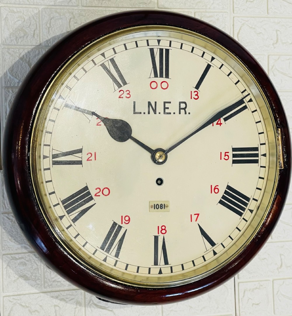 A circular LNER fusee railway wall clock featuring a dark mahogany wooden surround, brass bezel, cream dial with black Roman numerals and red 24-hour numbers. View V